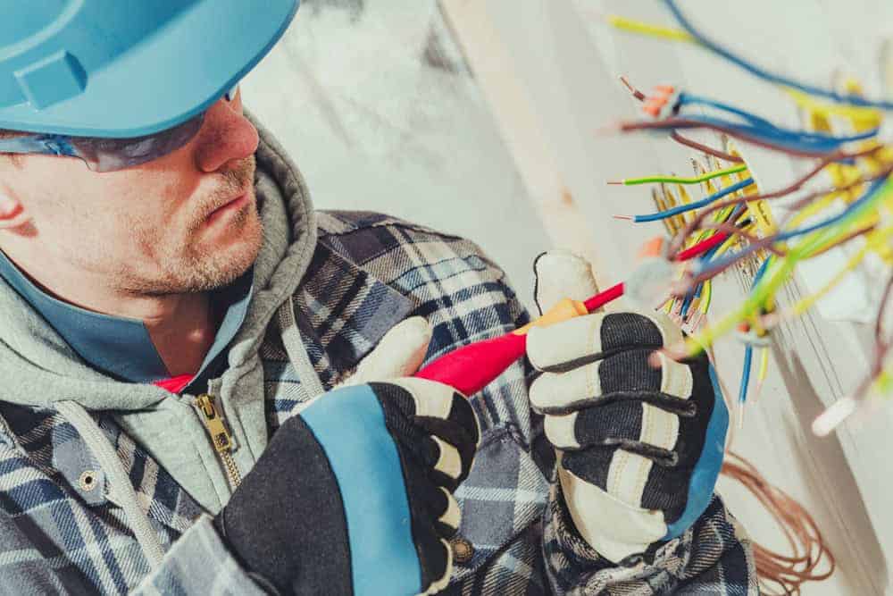 A construction worker, resembling a skilled residential electrician, dons a blue hard hat, safety glasses, and gloves as he expertly cuts electrical wires with red pliers. His focus is unwavering against a backdrop of exposed wiring in a wall, indicative of comprehensive electrical services underway.