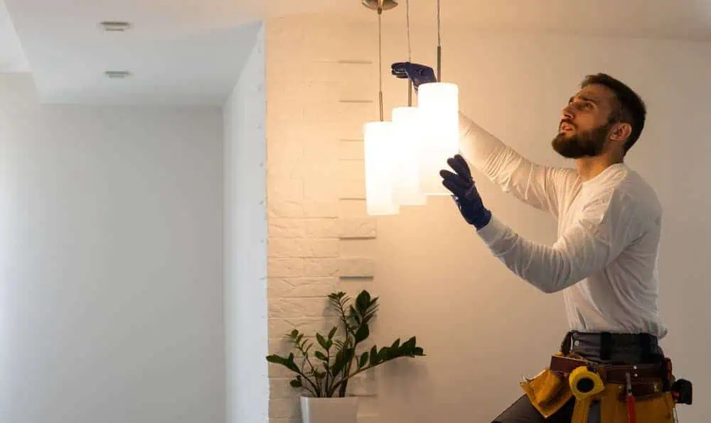 A residential electrician wearing work gloves and a tool belt is adjusting a hanging light fixture in a modern room. A white brick wall and a potted plant complete the scene, showcasing professional electrical services with precision and style.