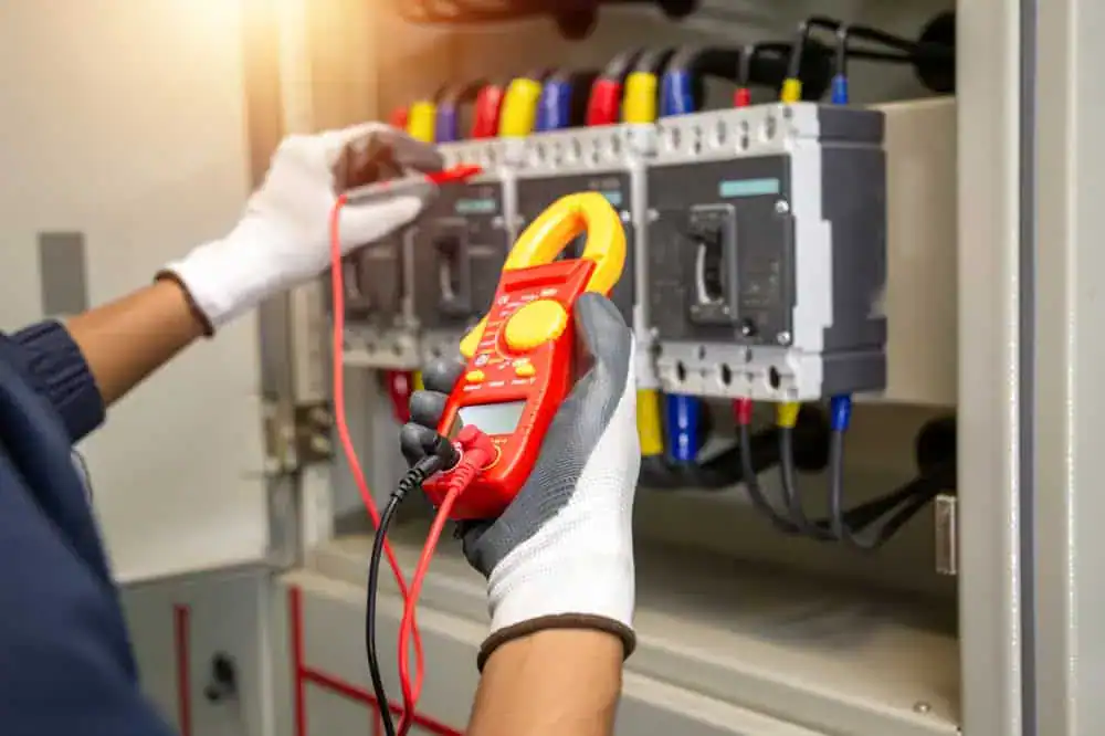 A residential electrician, wearing gloves, uses a red digital multimeter to test electrical components inside a panel. Several wires and switches are visible in the panel, with the individual's hand holding the device close to the components.