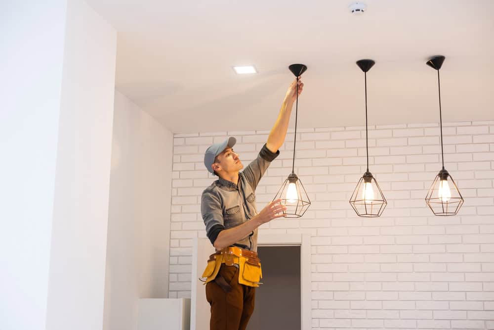 A residential electrician wearing a cap and tool belt adjusts one of the three hanging pendant lights in a room with white brick walls. The illuminated lights have wire cage designs, and the ceiling features a smoke detector and a recessed light, showcasing expert electrical services.