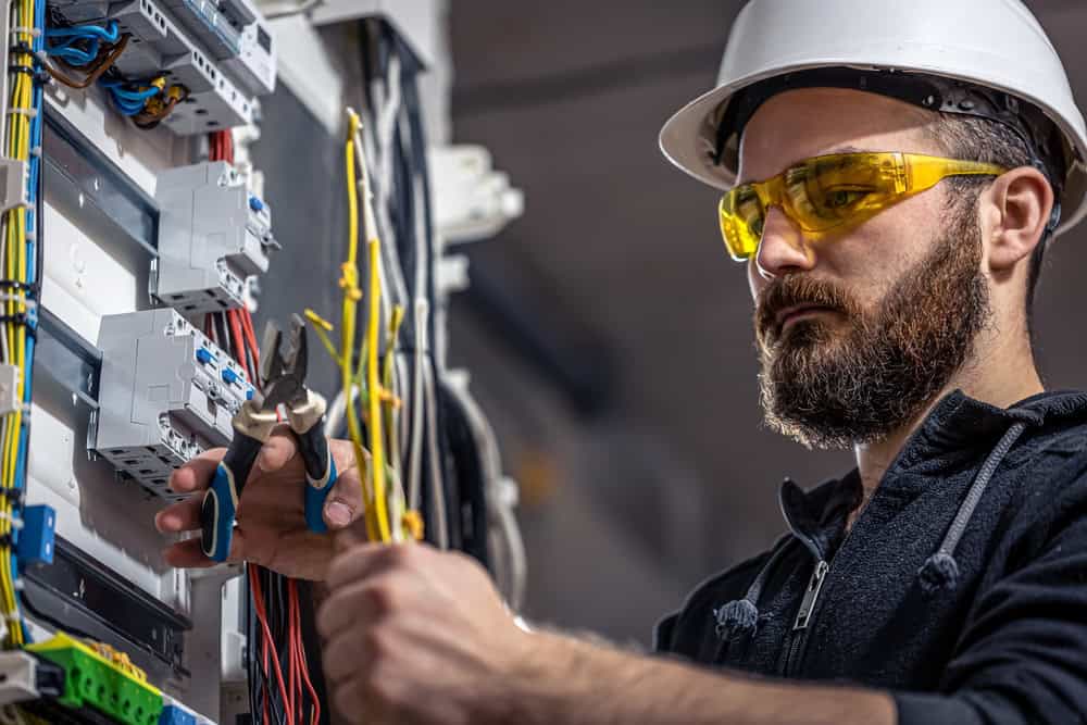The commercial electrician, wearing a white hard hat and yellow safety glasses, skillfully uses pliers to work on wiring in an electrical panel. He is focused on connecting the organized, colorful wires as part of his expert electrical services.