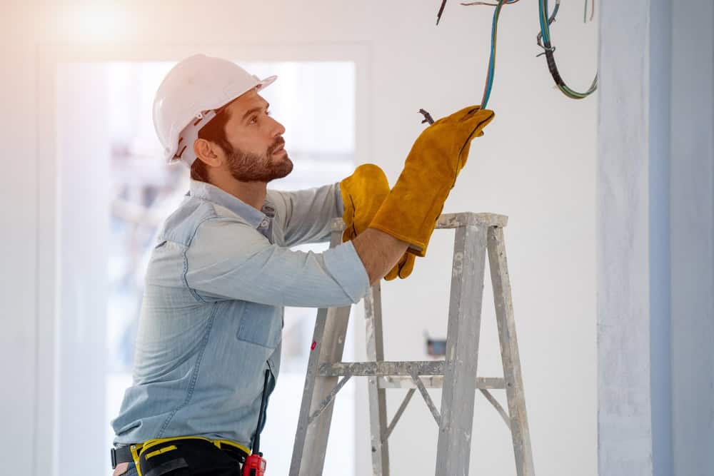 A construction worker wearing a white hard hat, yellow gloves, and a denim shirt stands on a ladder. He is busy with generator installation while handling exposed wires on the ceiling. The room is bright and unfinished.