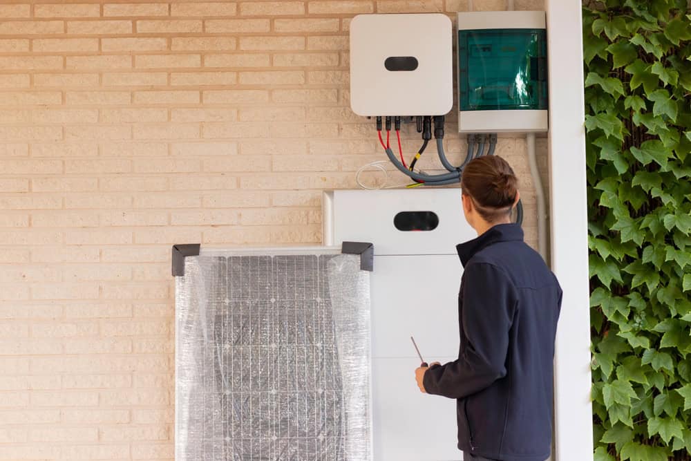 A residential electrician in a navy jacket, holding a screwdriver, stands in front of a wall-mounted energy storage system with wires, a solar panel, and battery pack. Green ivy grows on the adjacent wall.