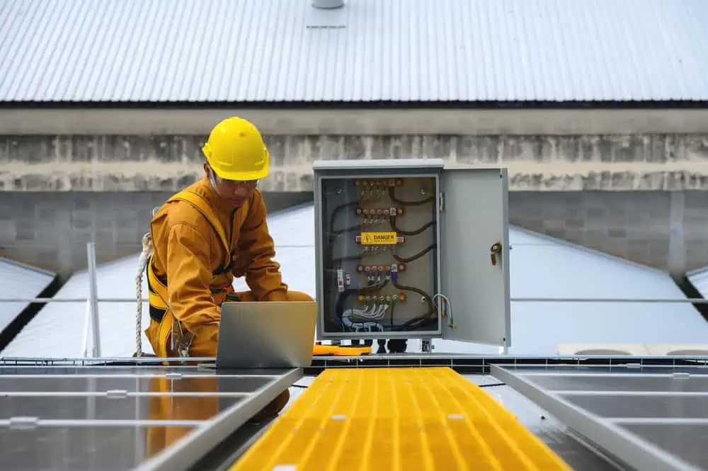A worker in safety gear, including a yellow helmet, meticulously inspects an open electrical panel on a rooftop. As a commercial electrician, they stand near solar panels for maintenance or installation purposes. The backdrop features a corrugated metal roof.