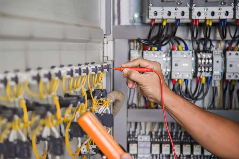 A commercial electrician is using a red probe to test wiring inside an electrical panel. The panel contains multiple yellow wires and various electronic components, including circuit breakers. The electrician is holding an orange multimeter to conduct the test efficiently.