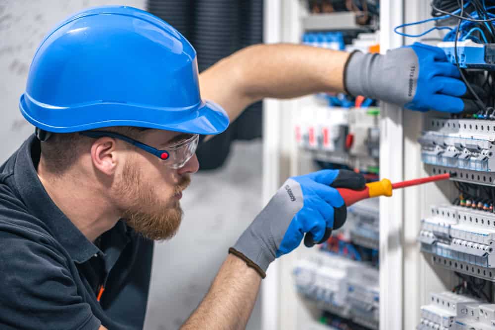 A residential electrician wearing a blue hard hat, safety glasses, and gloves works on an electrical panel. He skillfully uses an insulated screwdriver to adjust wires and components inside the cabinet, ensuring everything is safely and efficiently connected.