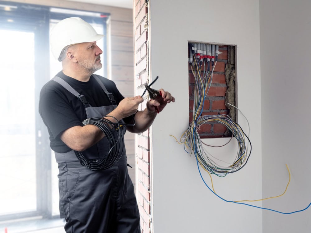 A commercial electrician in a hard hat and overalls holds a tool and a coil of electrical wire. He's standing beside a wall with exposed wiring, working near an unfinished area, with natural light streaming through large windows in the background.