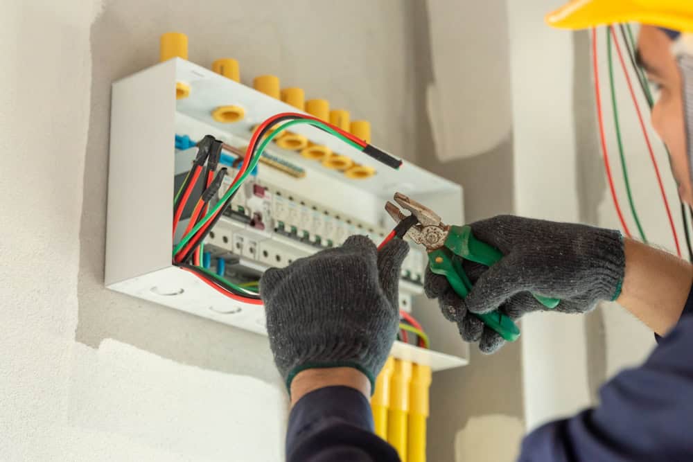 An electrician wearing gloves and a hard hat works on an electrical panel, expertly using pliers to handle wires. This commercial electrician ensures the colorful wires and circuit breakers are safely mounted, highlighting top-notch electrical services.