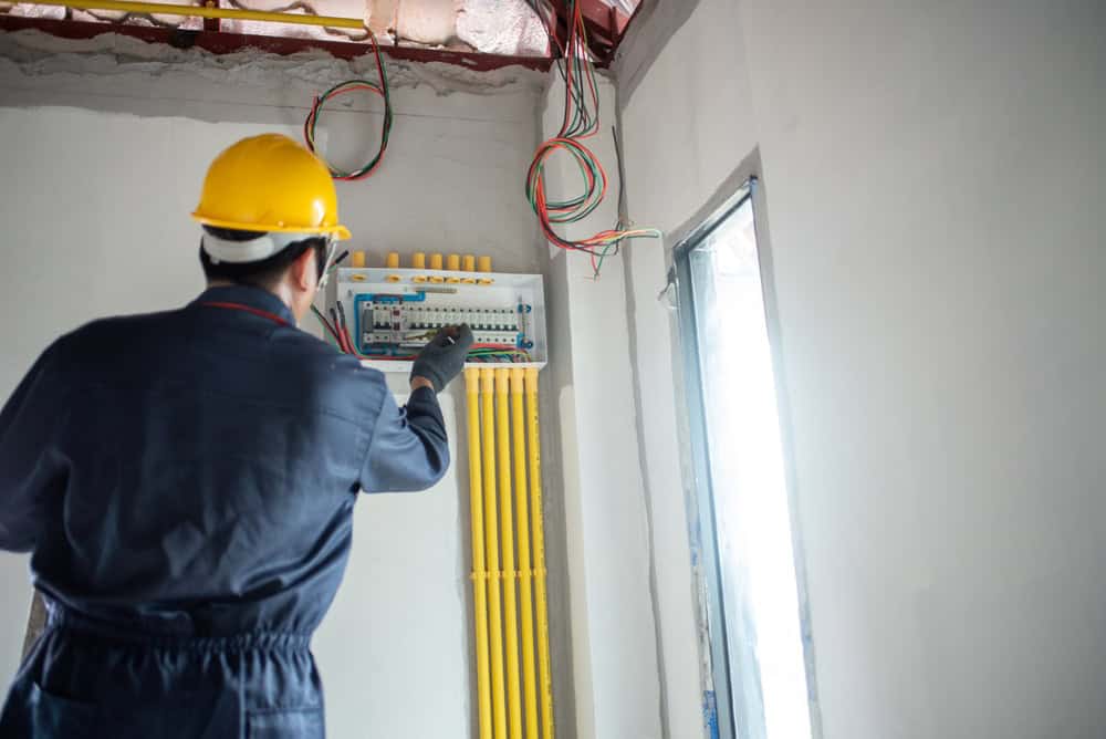 An electrician wearing a yellow hard hat and dark work clothes is adjusting an electrical panel on a white wall. As he provides essential electrical services, yellow cables run vertically below the panel while exposed wires are visible above. Natural light streams in from a nearby window.