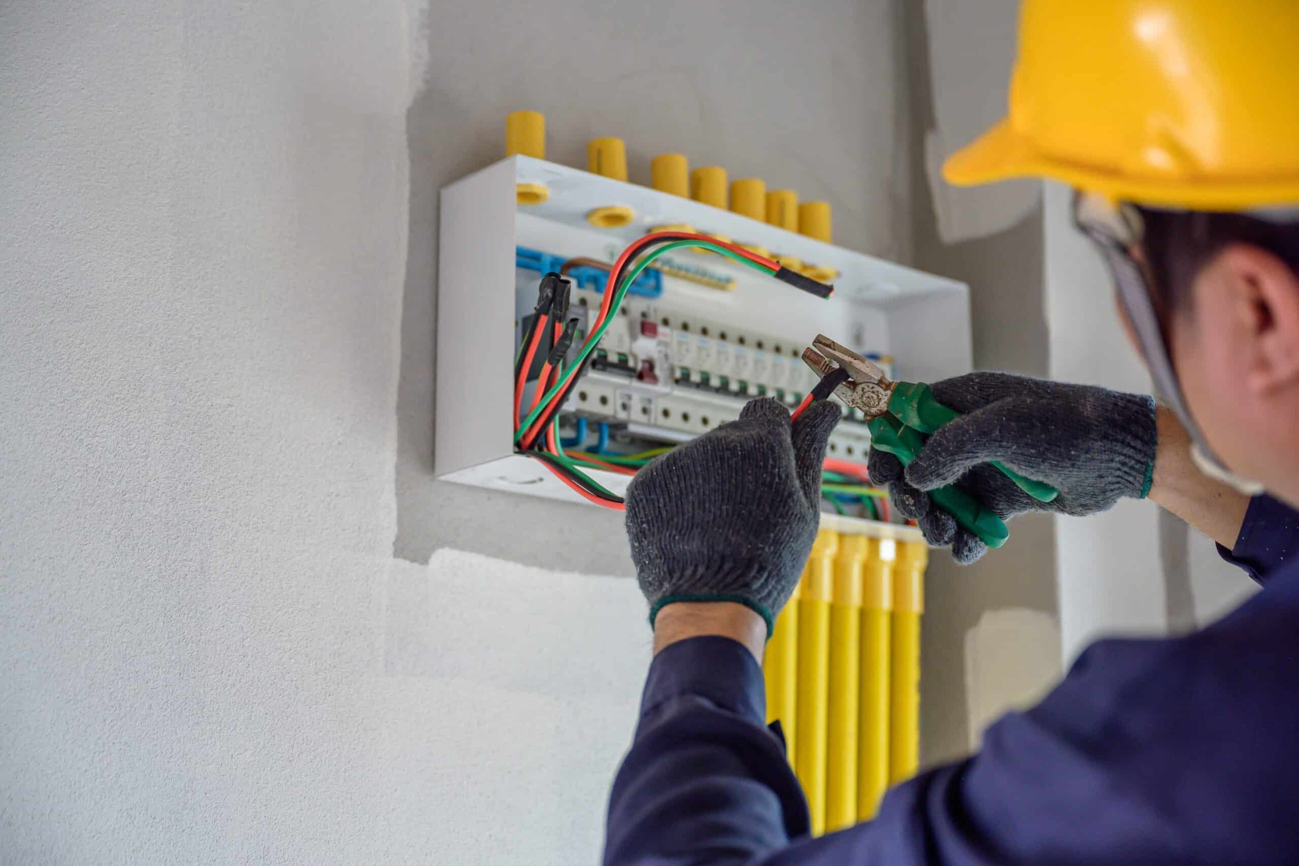 A person wearing a yellow hard hat and protective gloves is using pliers to work on a wiring panel with colorful cables in a white enclosure against a wall, showcasing the skills of a commercial electrician.
