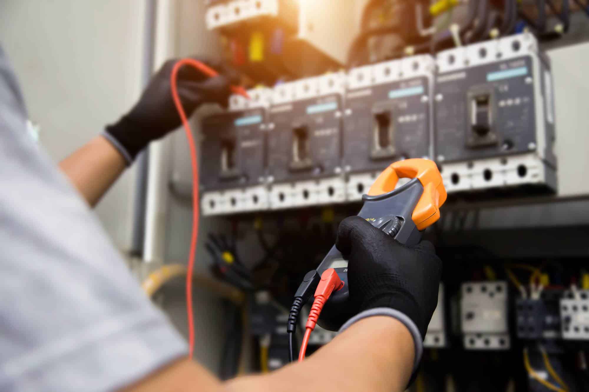 A person wearing black gloves uses a digital clamp meter to measure electrical current in a circuit breaker panel with multiple switches and wires.