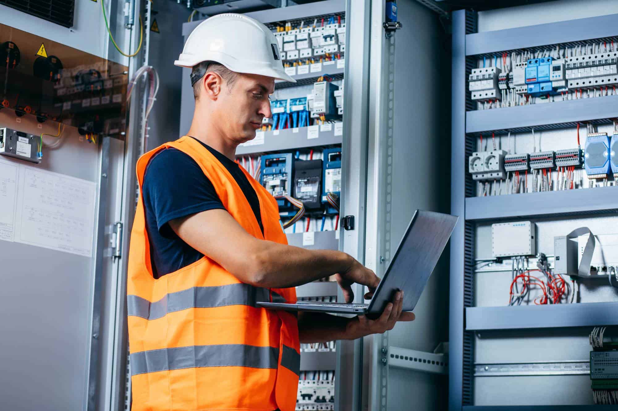 A worker in a white hard hat and orange safety vest uses a laptop while standing in front of open electrical panels filled with wiring and components.