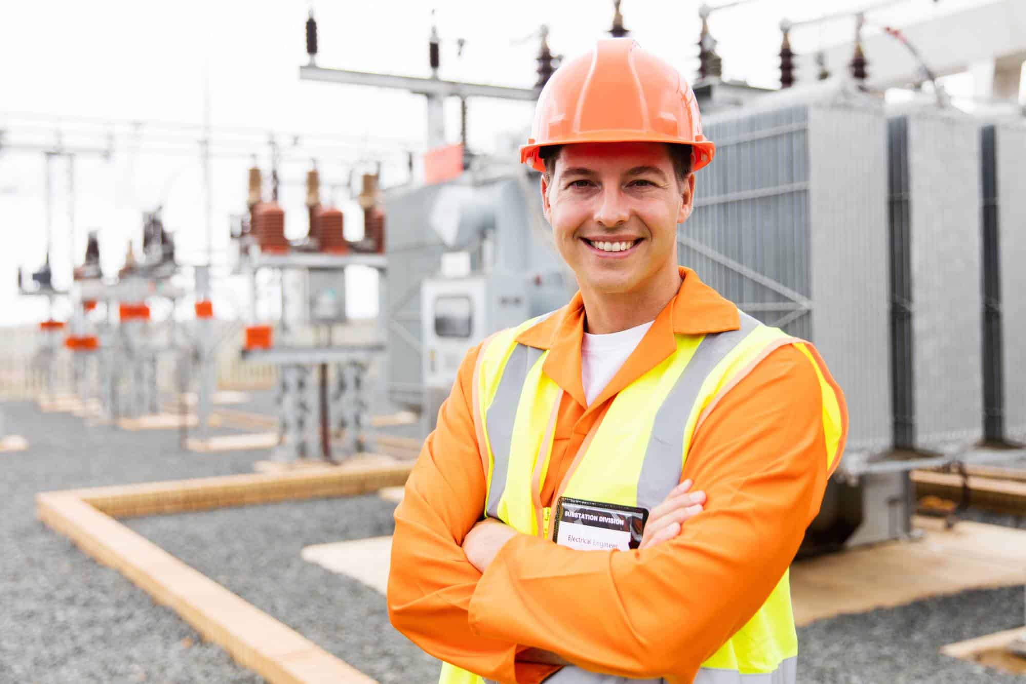 A smiling engineer in an orange safety jacket and hard hat stands with arms crossed at an outdoor electrical substation, with transformers and electrical equipment in the background.