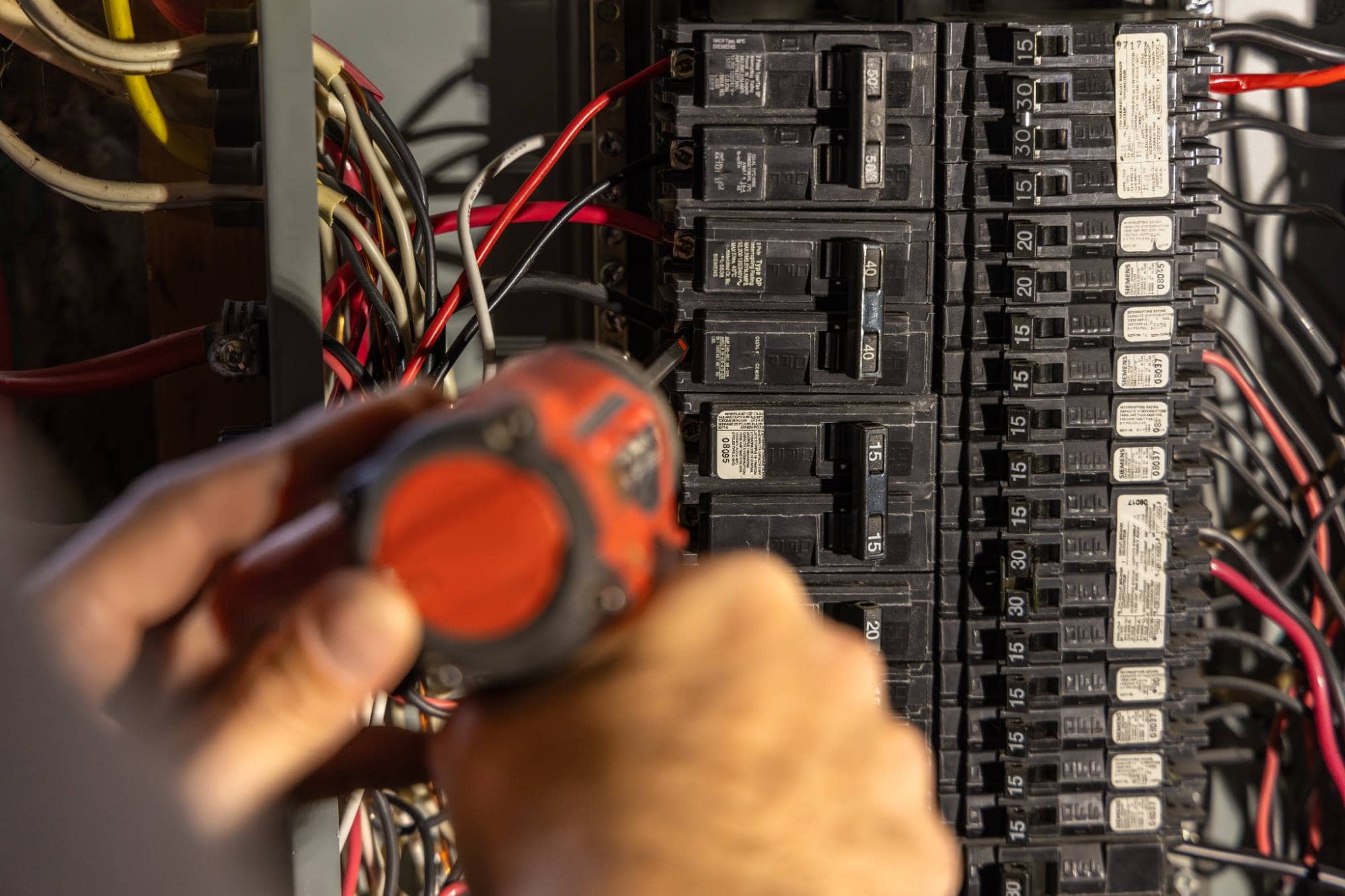 A person uses a red cordless drill to work on an open electrical breaker panel filled with black switches and various colored wires.