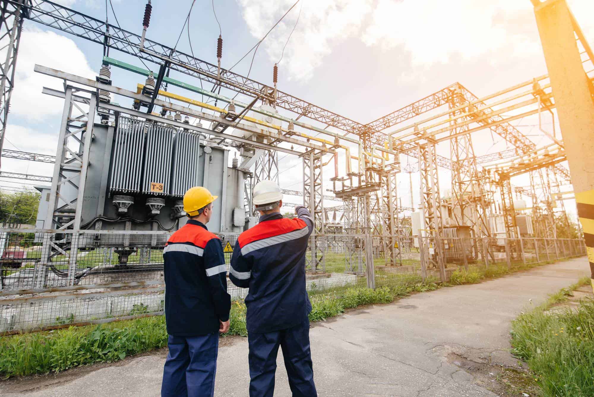 Two workers in protective gear and helmets stand near high-voltage electrical equipment at an outdoor power substation, inspecting the site under a bright, partly cloudy sky.