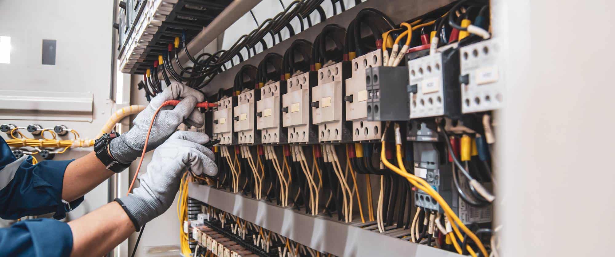 An electrician wearing gloves uses a probe tool to check wiring in an electrical control panel filled with multiple relays, switches, and connected yellow and black wires.