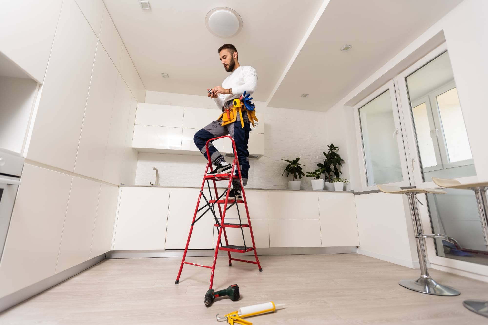 A handyman stands on a red step ladder in a modern white kitchen, wearing a tool belt and working with tools. Tools are placed on the floor nearby.