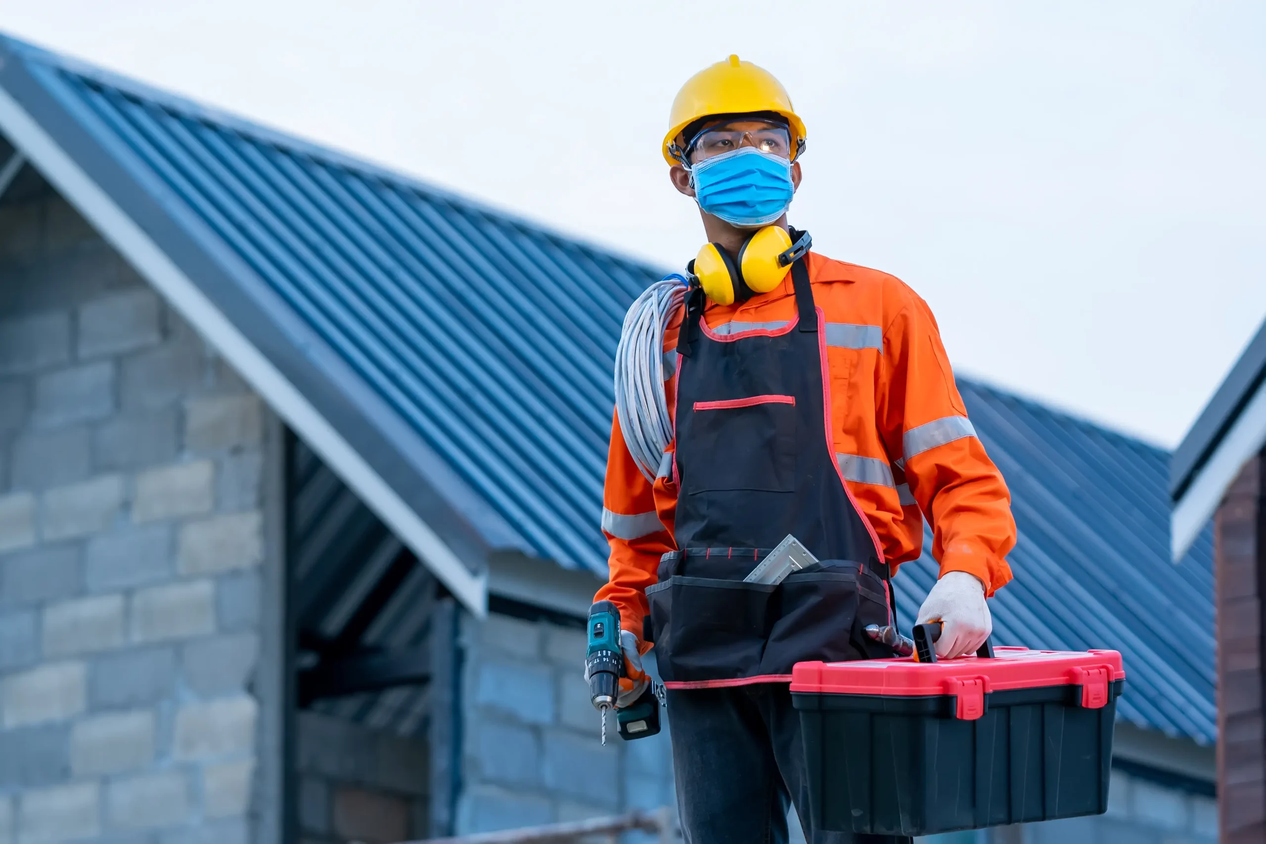 A construction worker wearing a hard hat, face mask, and safety gear holds a toolbox, a power drill, and coiled cable, standing in front of a building under construction.
