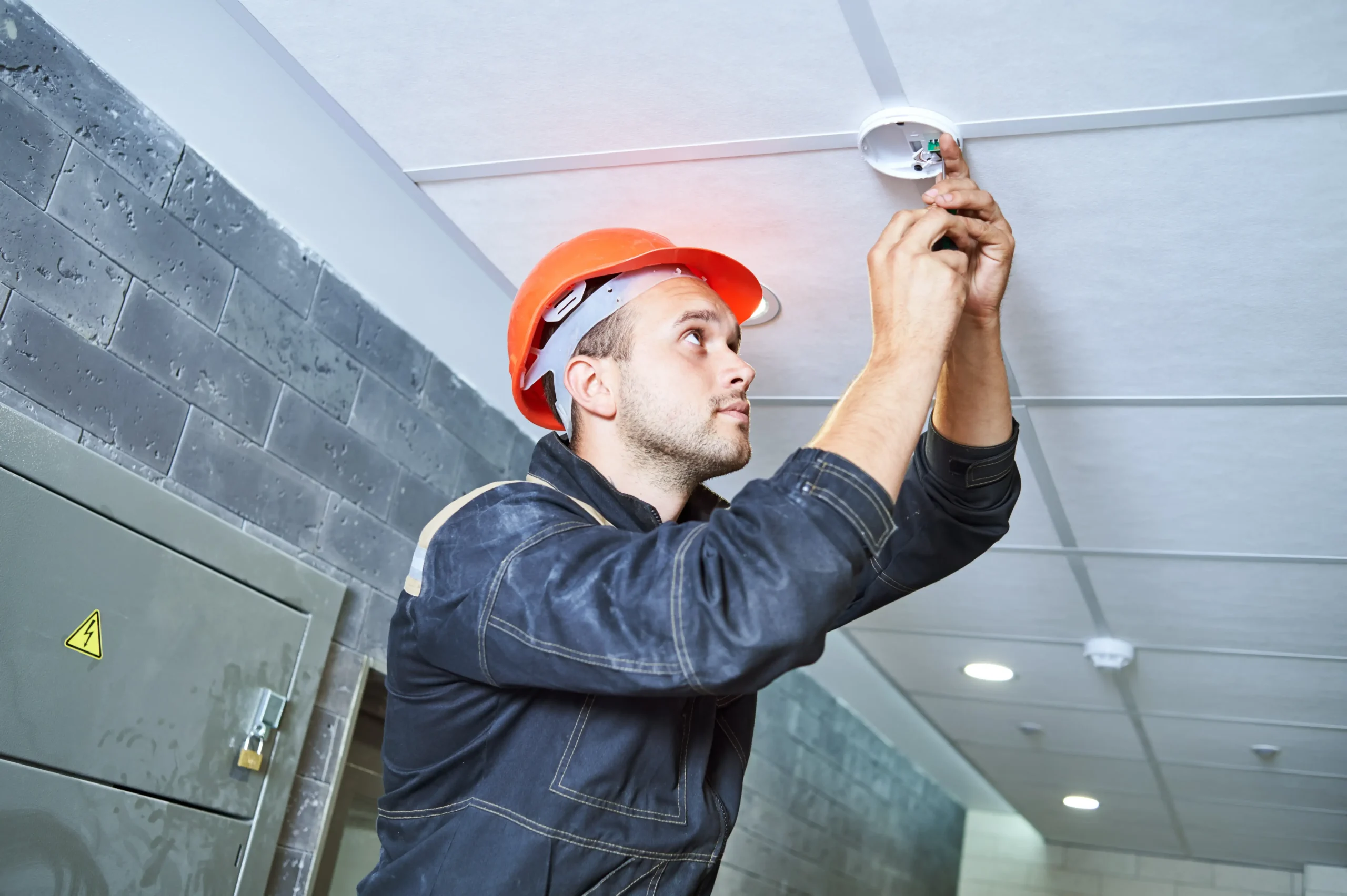 A worker wearing a red hard hat and dark coveralls installs or repairs a smoke detector on a white tiled ceiling in a hallway with gray brick walls.
